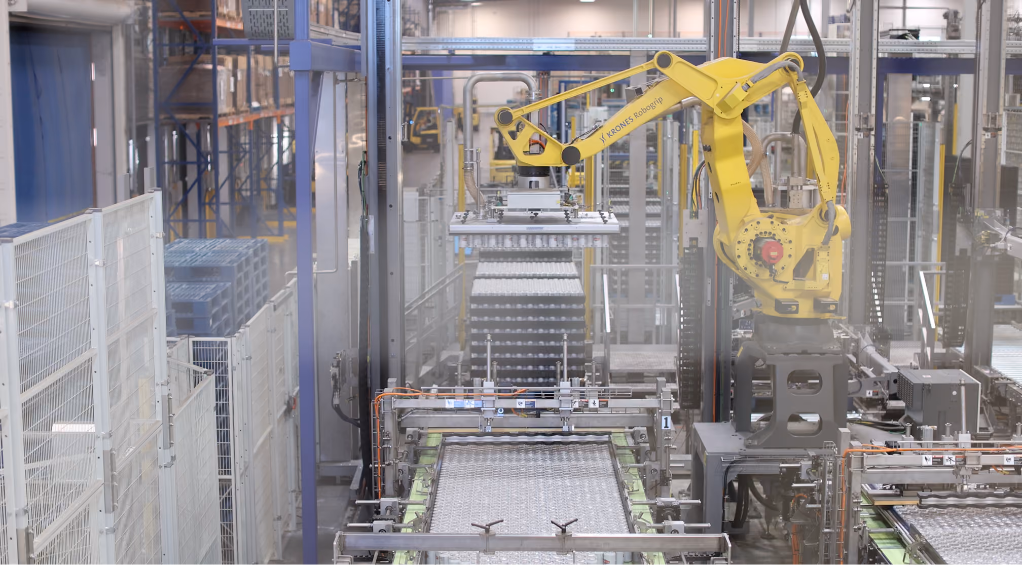 Yellow robotic arm labeled KRONES Robogrip in a factory moving trays of cans on a conveyor belt.