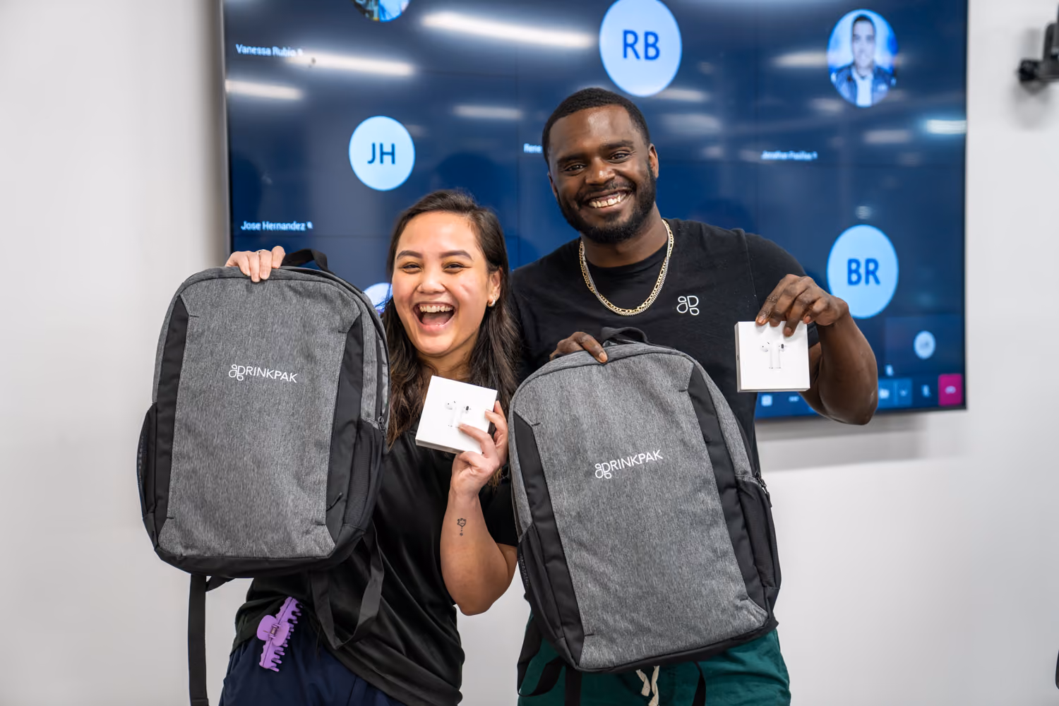Two smiling people holding grey DrinkPak backpacks and white wireless earbud boxes in an office.