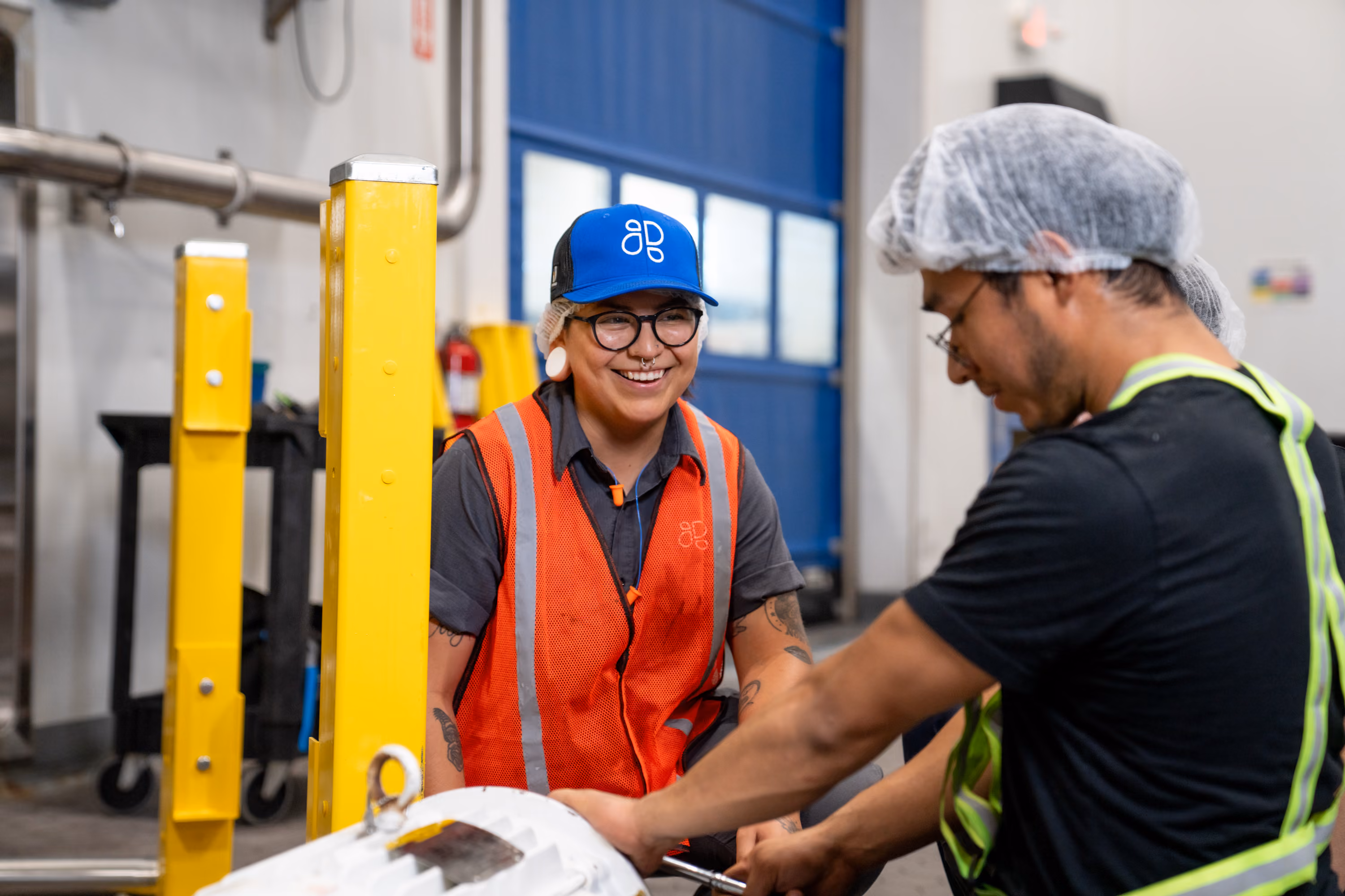 Two factory workers in safety gear collaborating on a machine part inside an industrial space.
