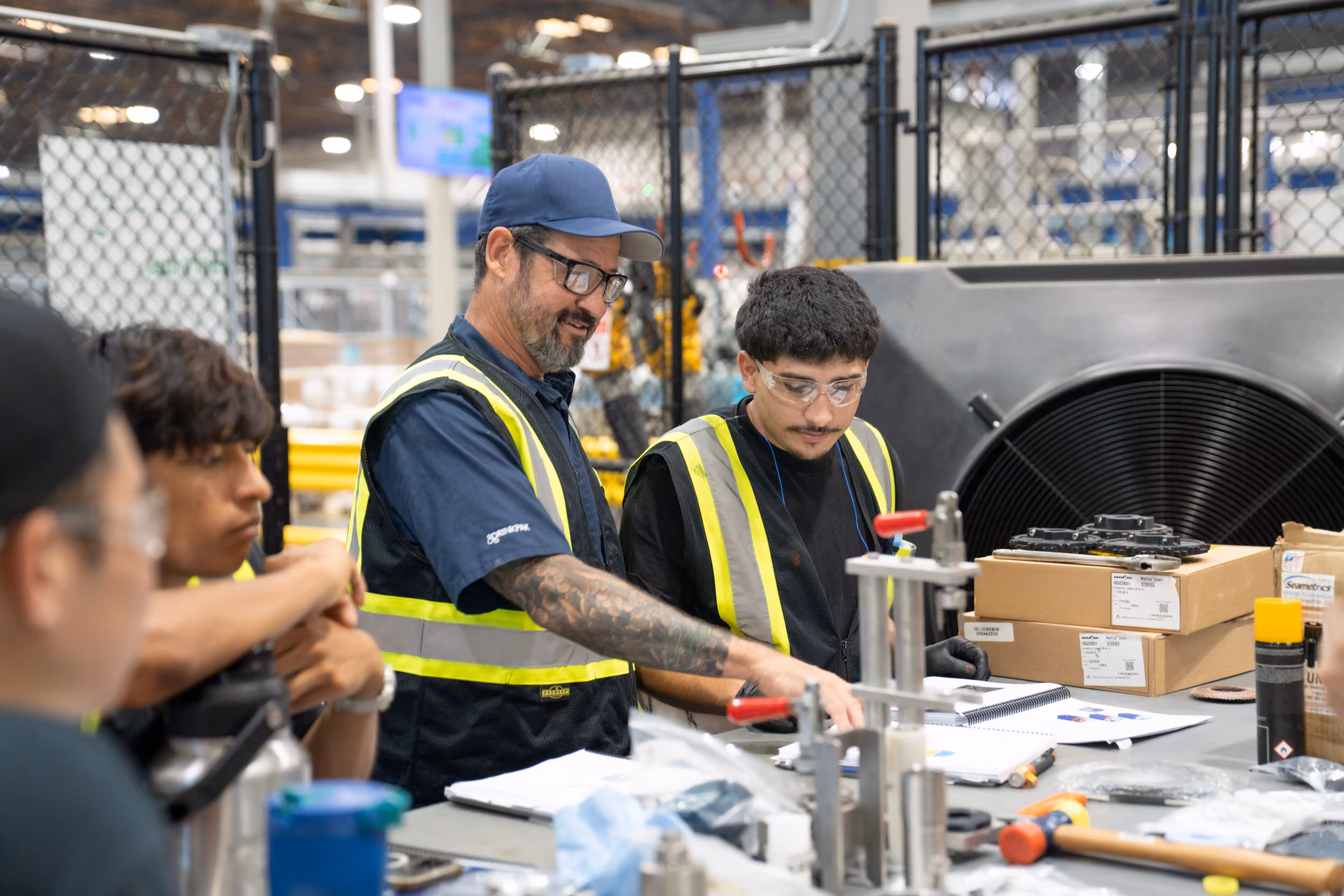 Workers in safety vests discuss documents at a factory workspace with boxes and tools nearby.