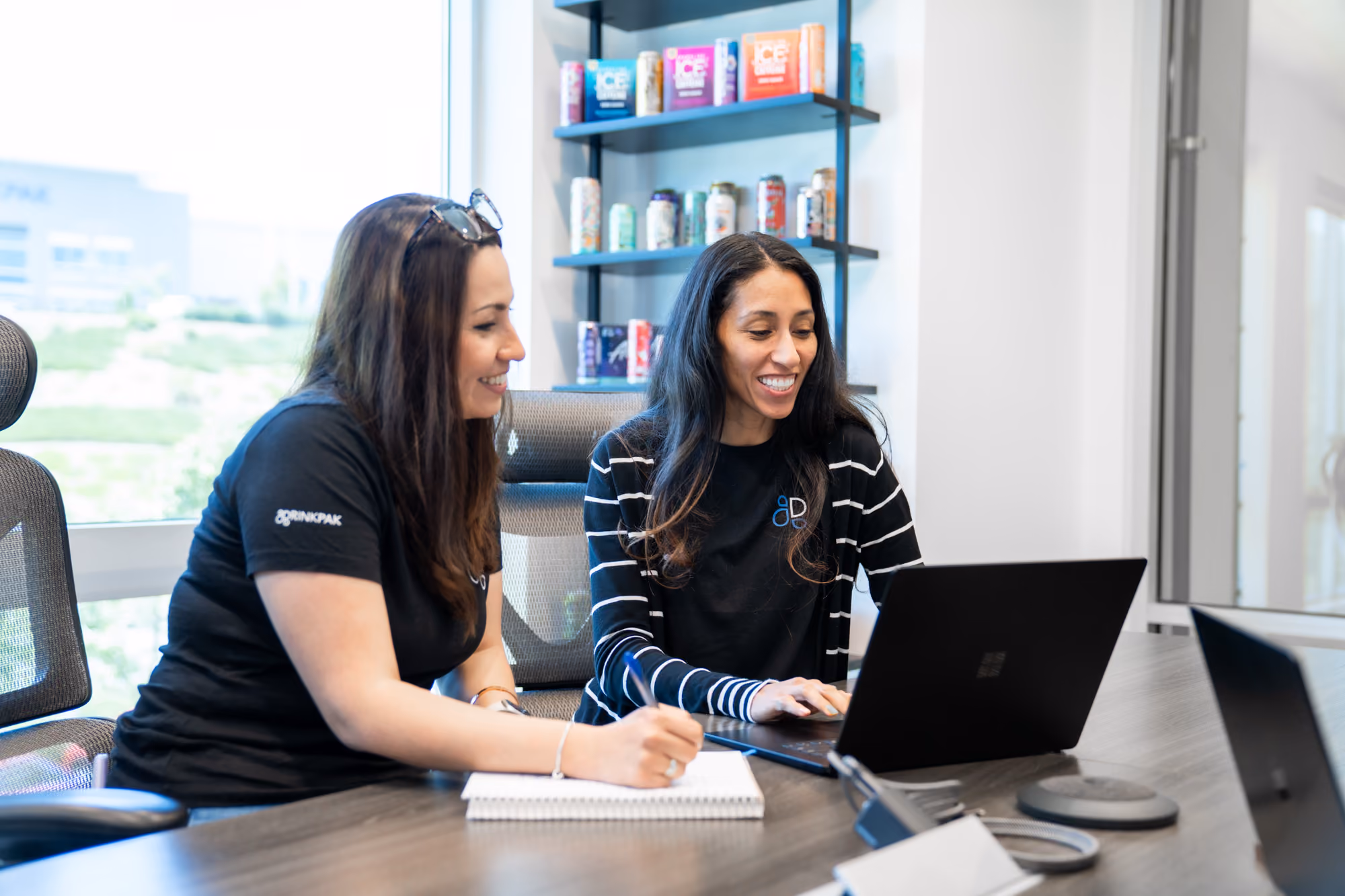 Two women working together at a table, one typing on a laptop, the other writing notes in a notebook.