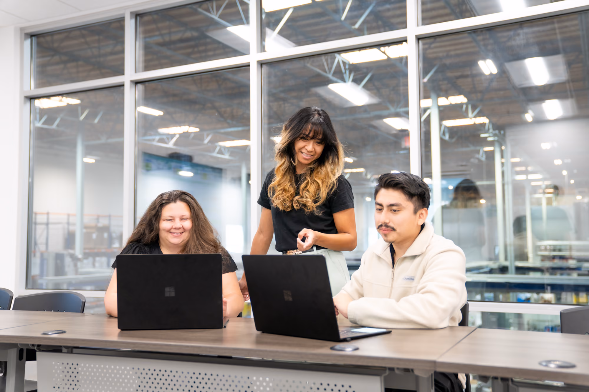 Three people working and collaborating on laptops at a table in an office with glass walls.