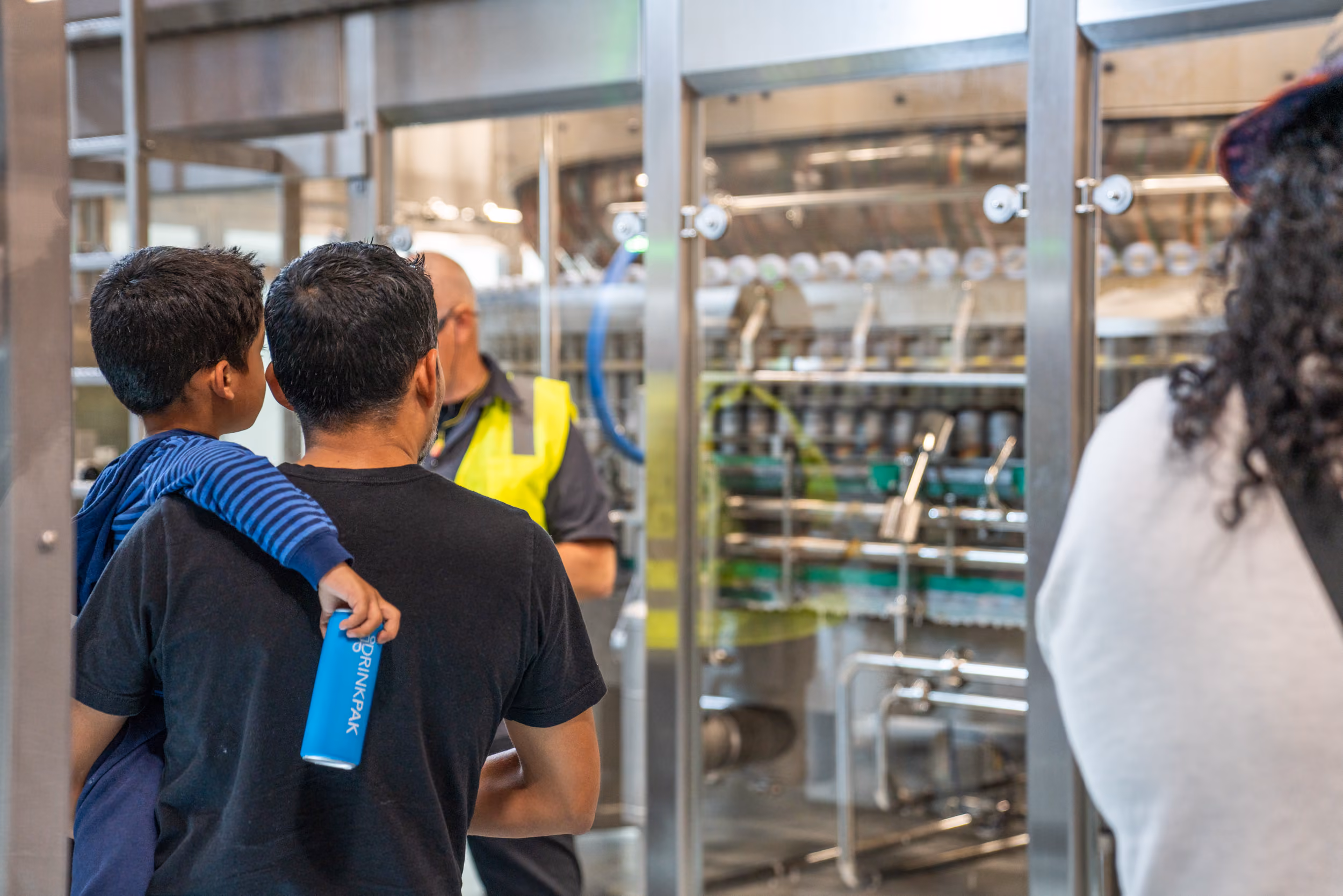 Man carrying a boy holding a blue Drinkpak bottle inside a factory with a worker in a yellow vest.