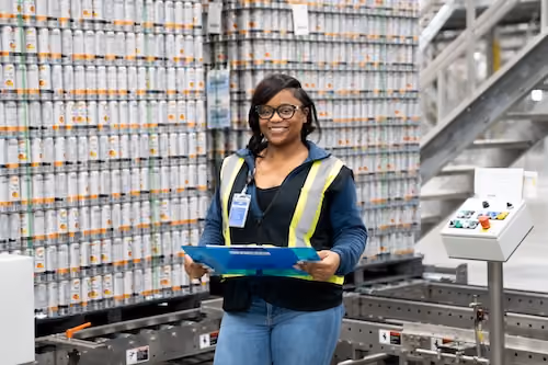 Smiling woman in safety vest holding clipboard in a warehouse with stacked cans behind her.