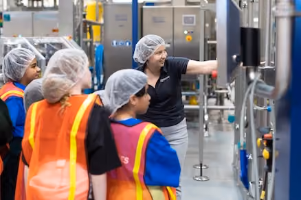 Group wearing hairnets and safety vests in a factory observing a woman pointing at machinery controls.