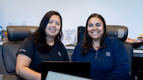 Two smiling women sitting next to each other at a desk with laptops in an office setting.