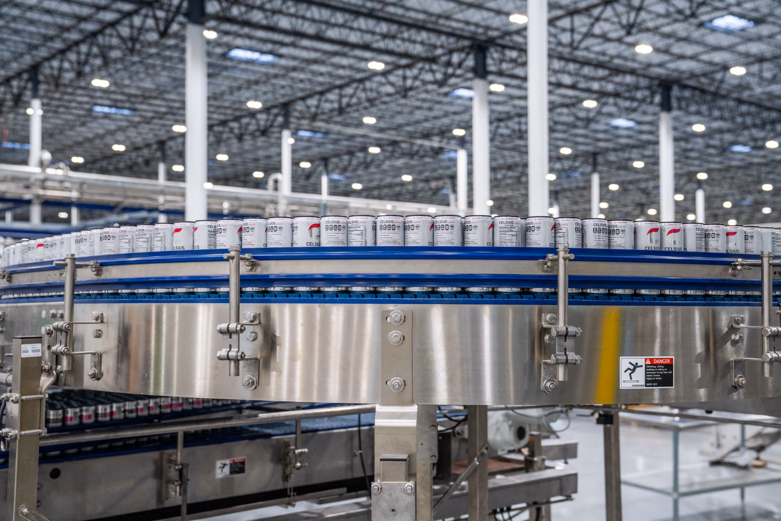 Rows of Celsius beverage cans on a conveyor belt inside a large, well-lit industrial factory.