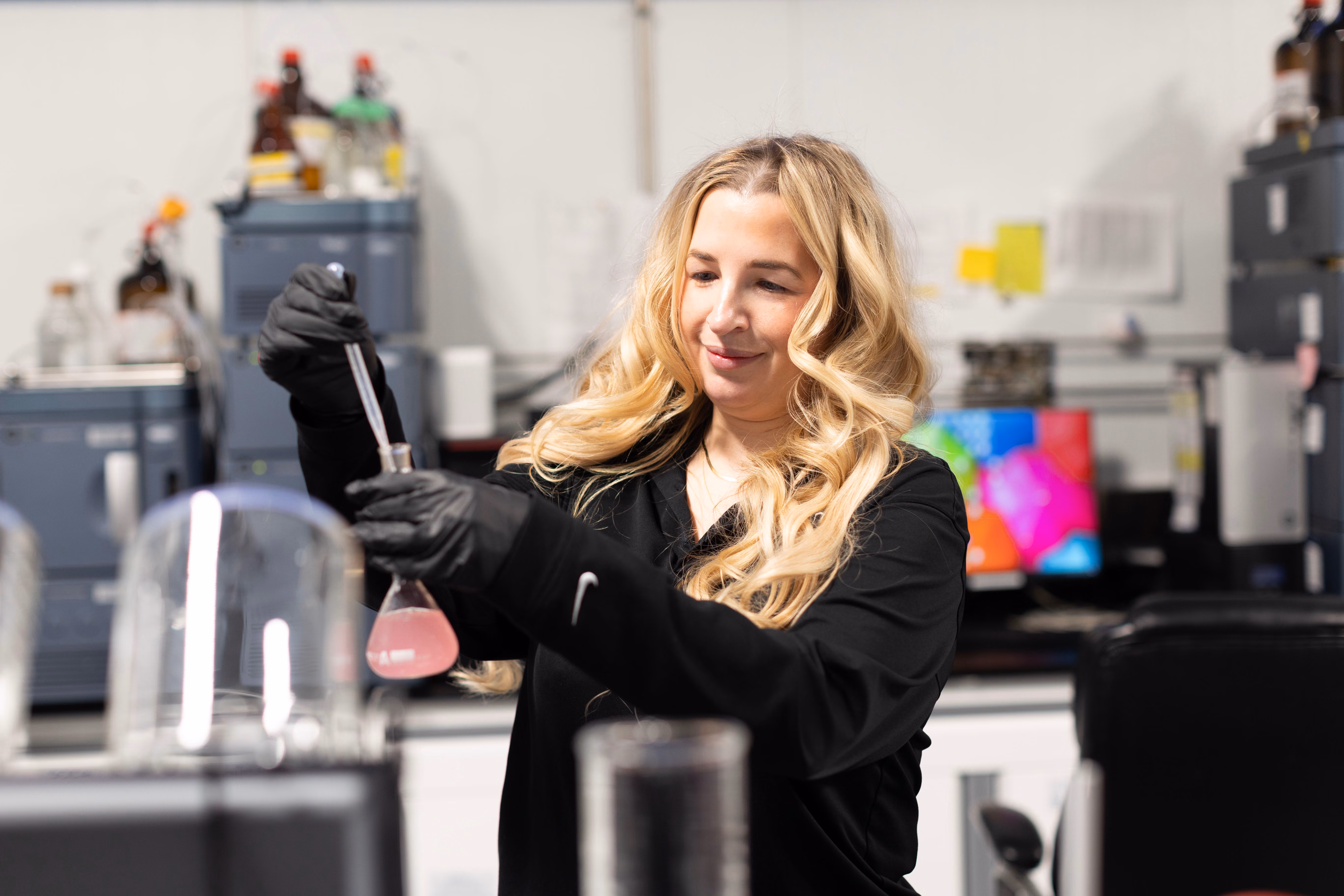 Scientist with long blonde hair wearing black gloves works with a pink liquid in a flask in a lab.