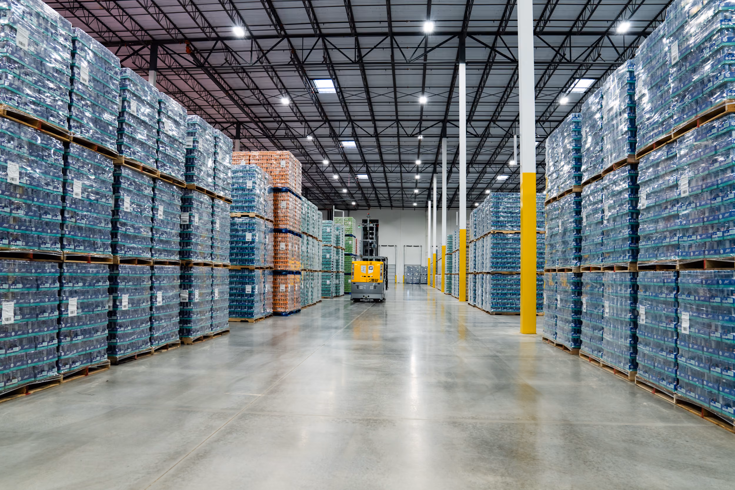 Spacious warehouse aisles lined with tall stacks of palletized goods and a yellow forklift in the center.