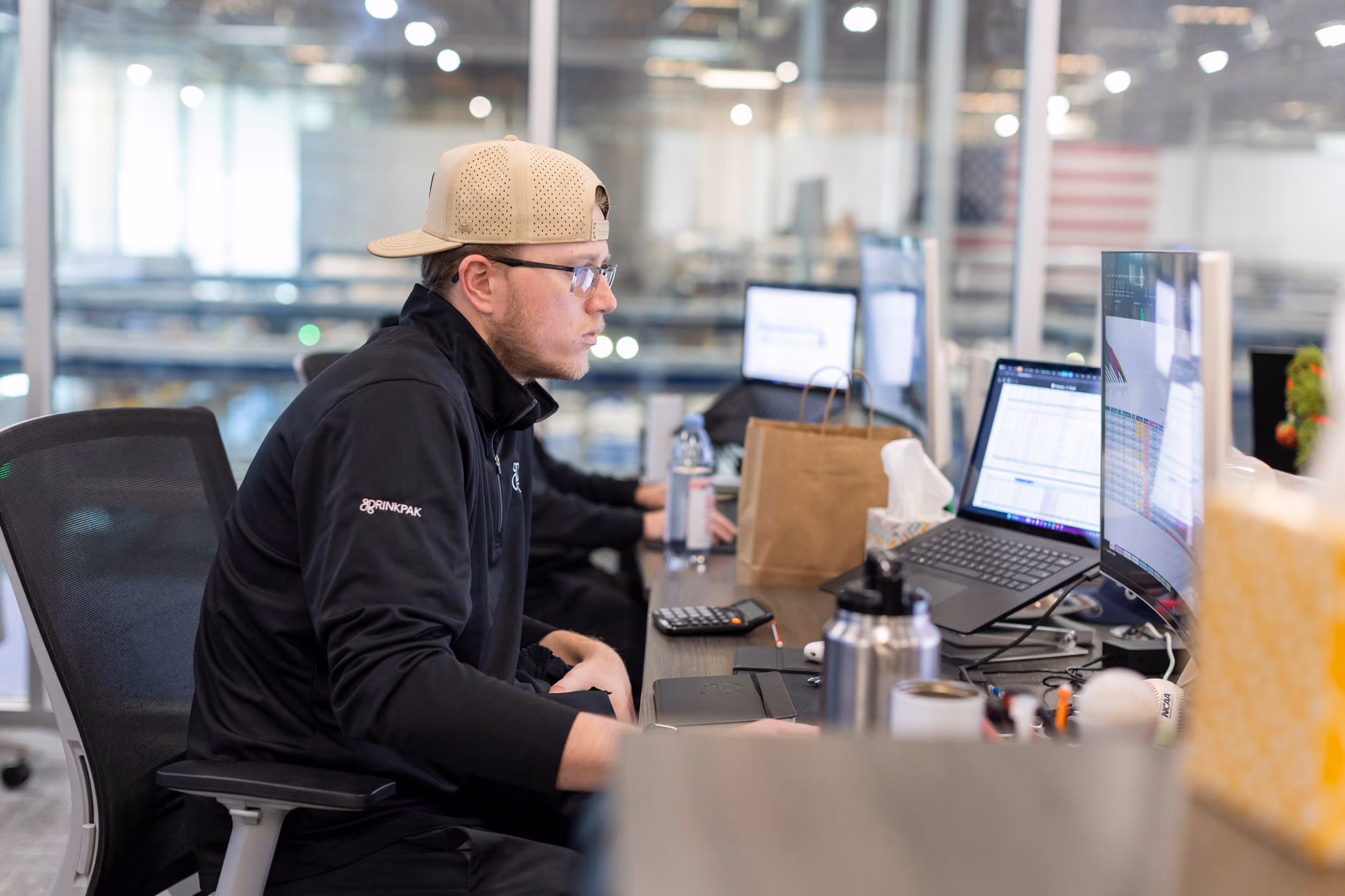 Man in a beige cap and glasses working at a desk with multiple computer screens displaying data.