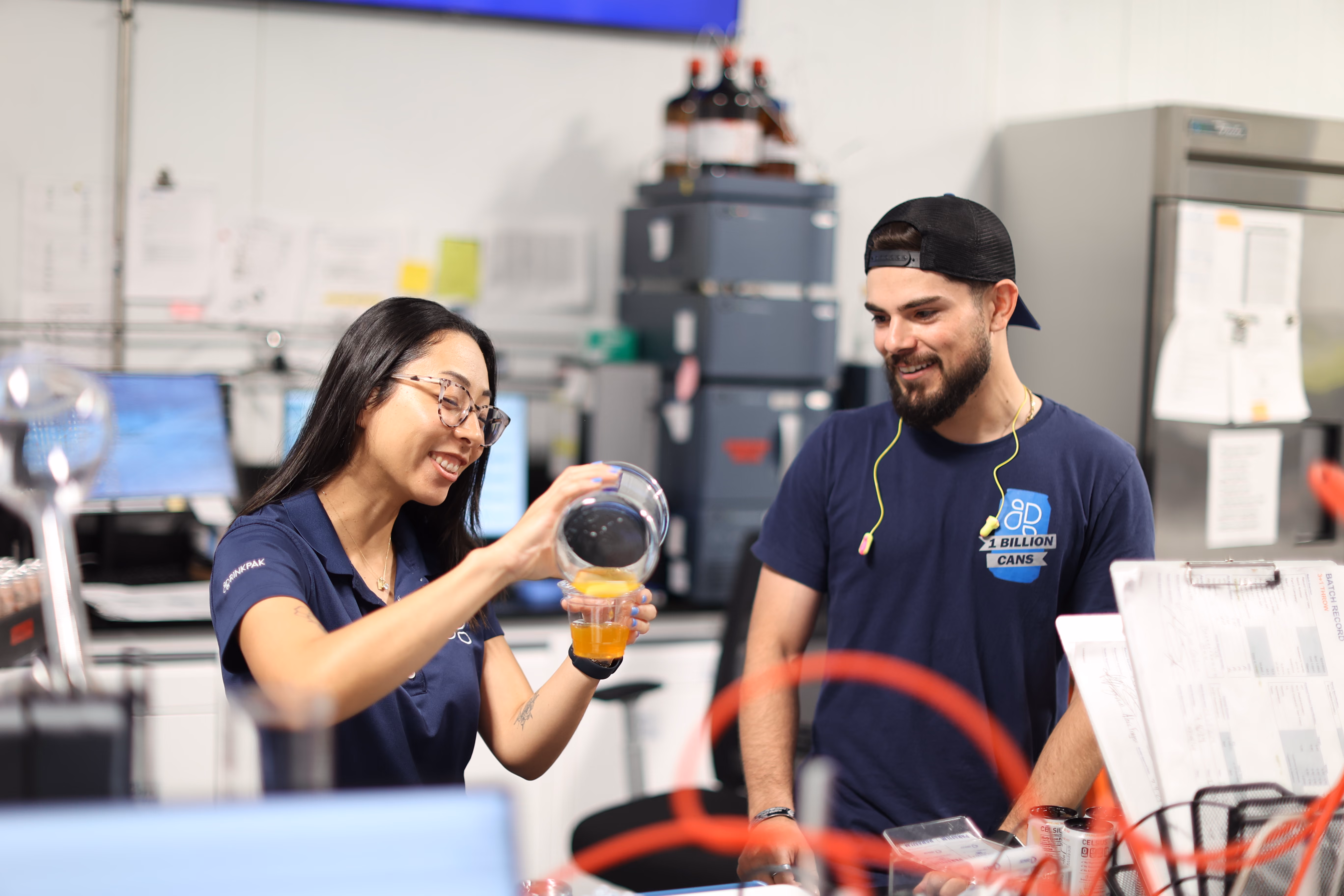 Two people in a lab or workspace, one pouring orange liquid into a cup while the other watches smiling.