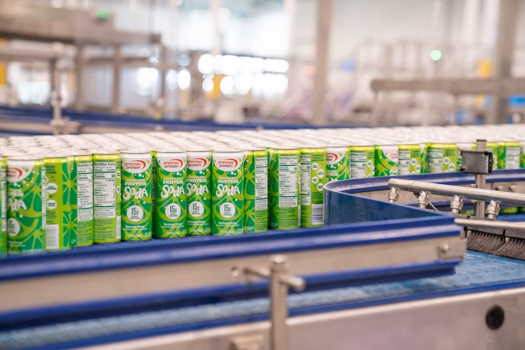 Cans of Premier Protein Soy Soda lined up on a blue factory conveyor belt.