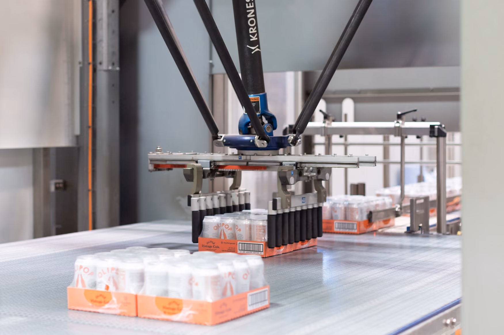 Robotic arm packaging multiple trays of canned beverages on a conveyor belt in a factory.