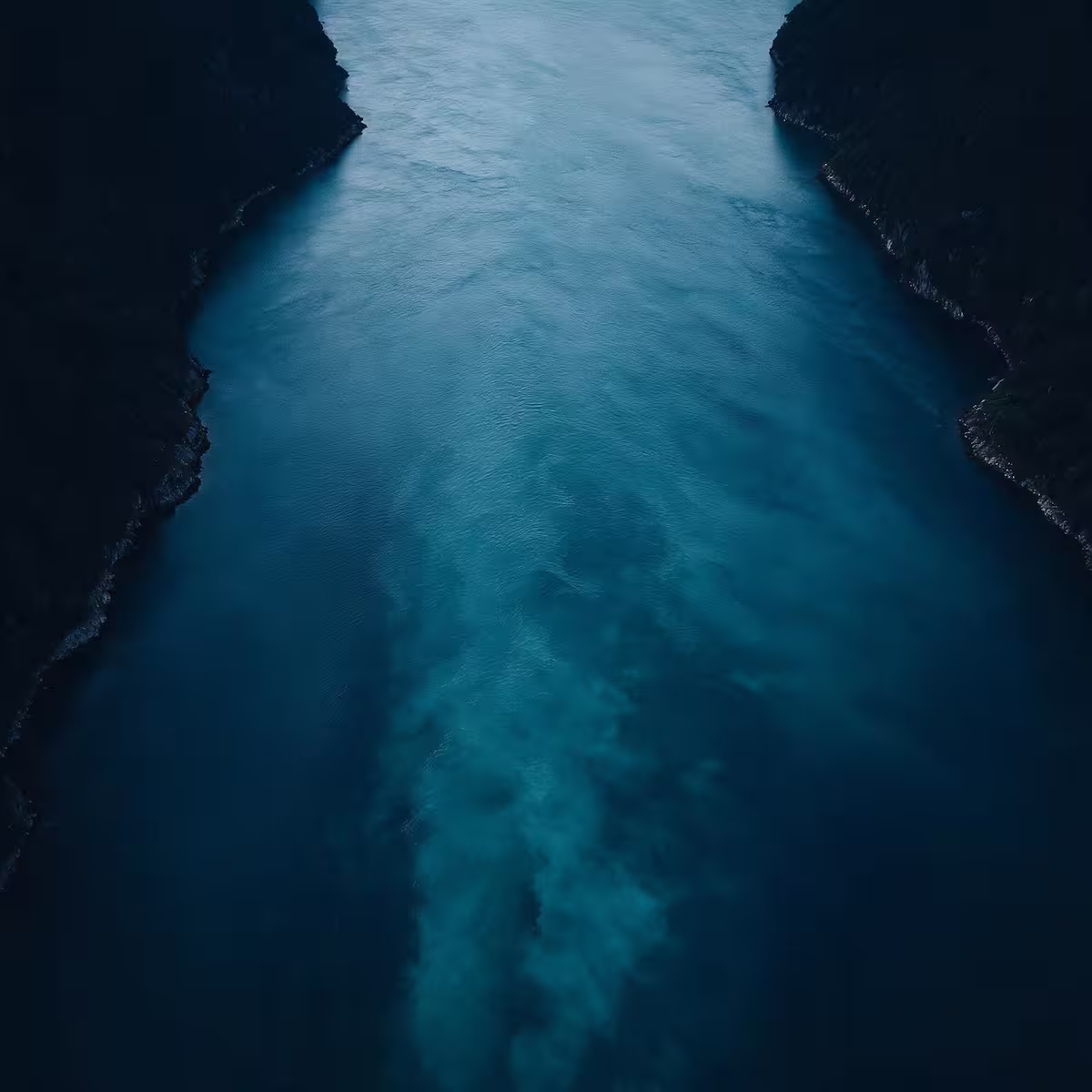 Oversiktsbilde av en smal fjord med mørke fjell på begge sider og blått vann i midten.