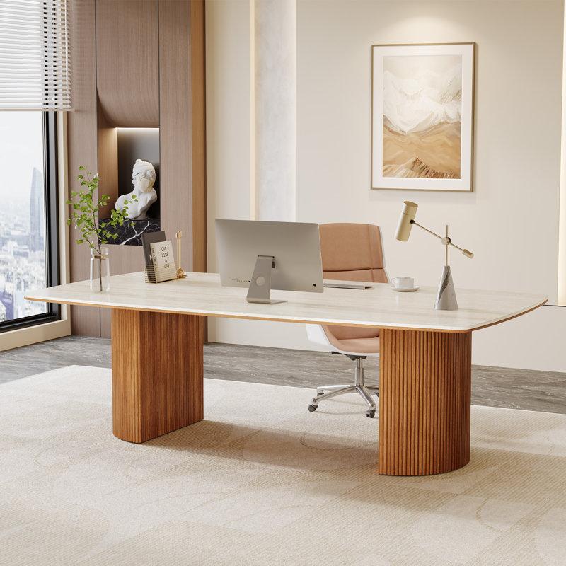 Modern minimal office space with a large light wood desk supported by two wooden pedestal legs, a beige office chair with wheels, a desktop computer, a gold desk lamp with a stone base, a small coffee cup, a calendar, and a glass vase with green leafy branches, against a light wall featuring an abstract framed artwork and a recessed niche with a white bust sculpture.