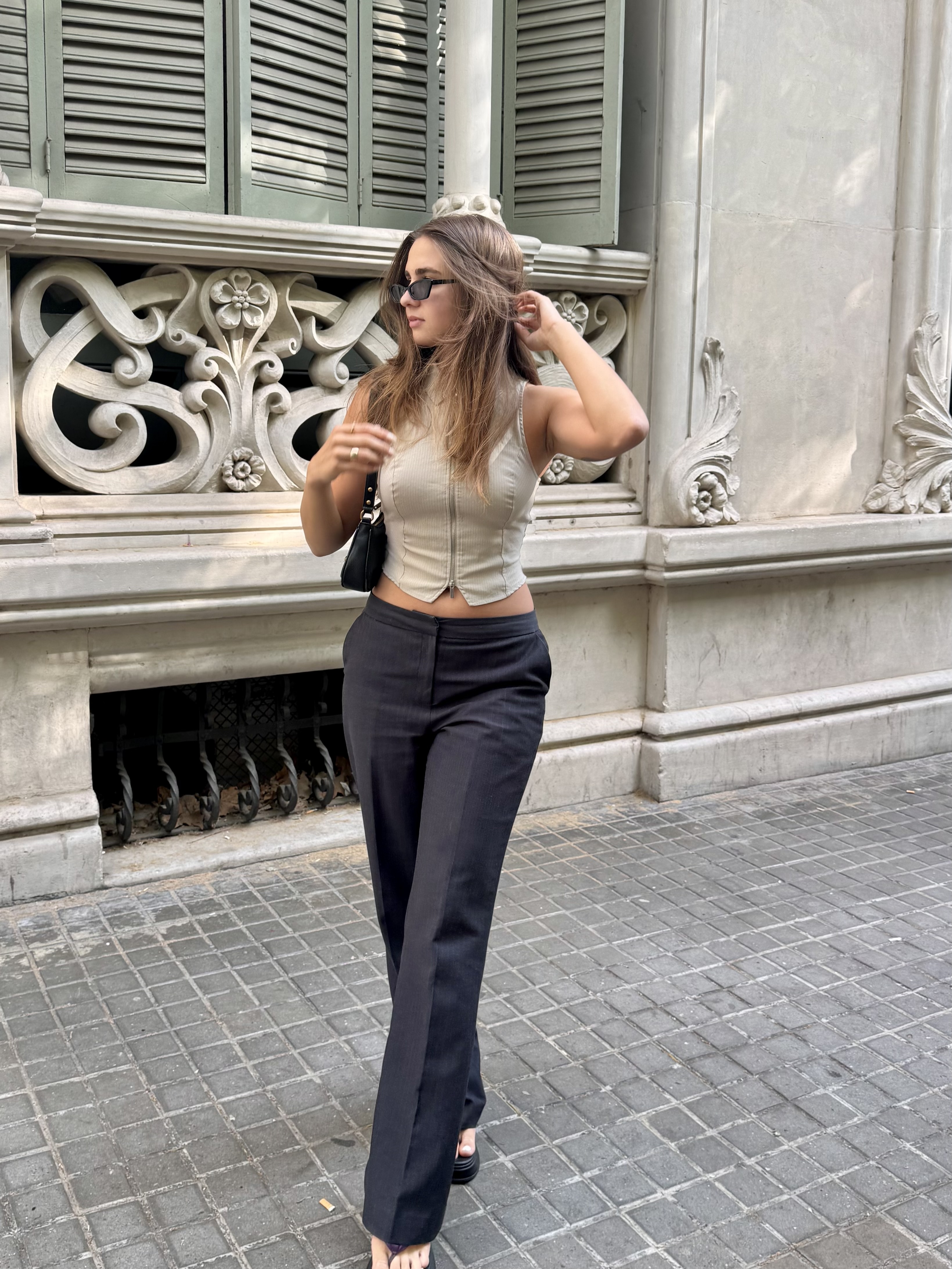 Woman with long hair wearing sunglasses, beige sleeveless top, black pants, and carrying a black purse.