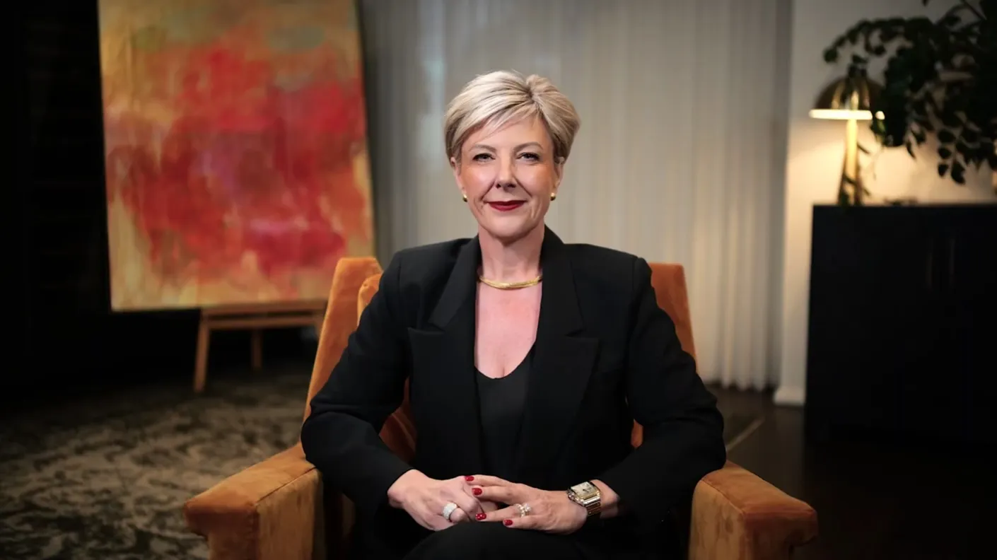 Smiling Nicole Moodly with short blond hair wearing black suit sitting in orange armchair indoors.