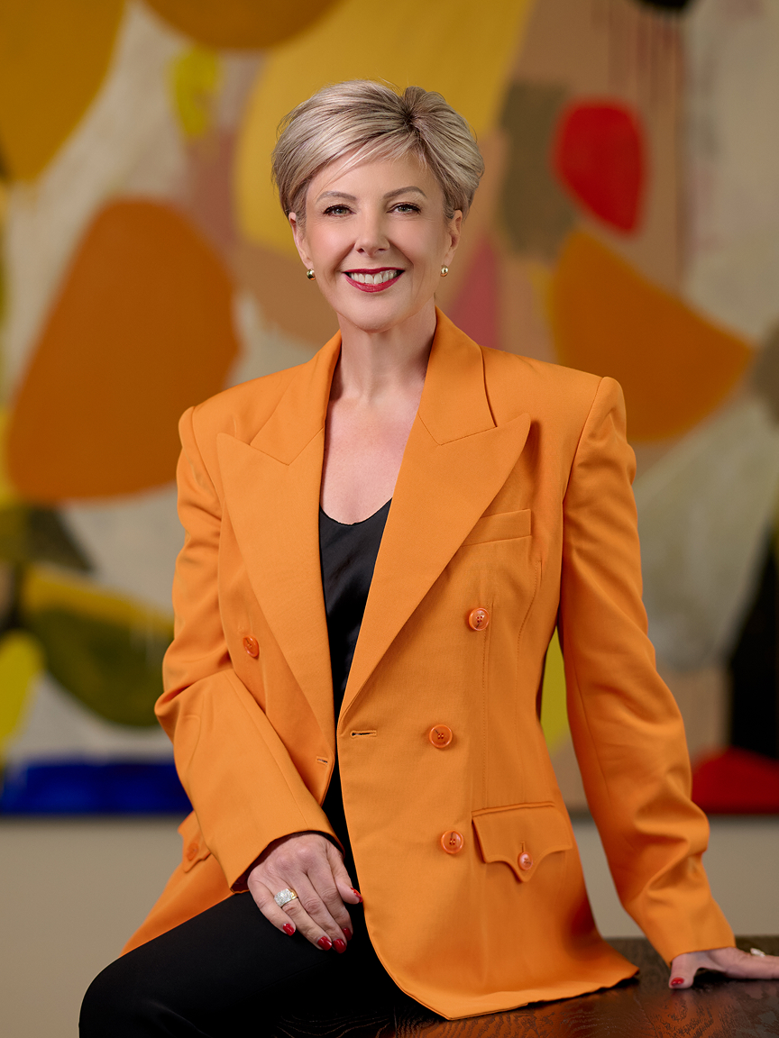 Smiling Nicole Moody with short blonde hair wearing an orange blazer sitting on a table with colorful abstract background.