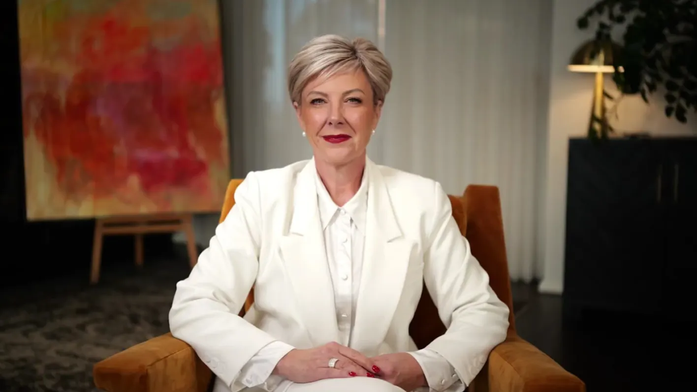 Nicole Moody with short blonde hair wearing a white blazer seated in a rust-colored chair indoors.