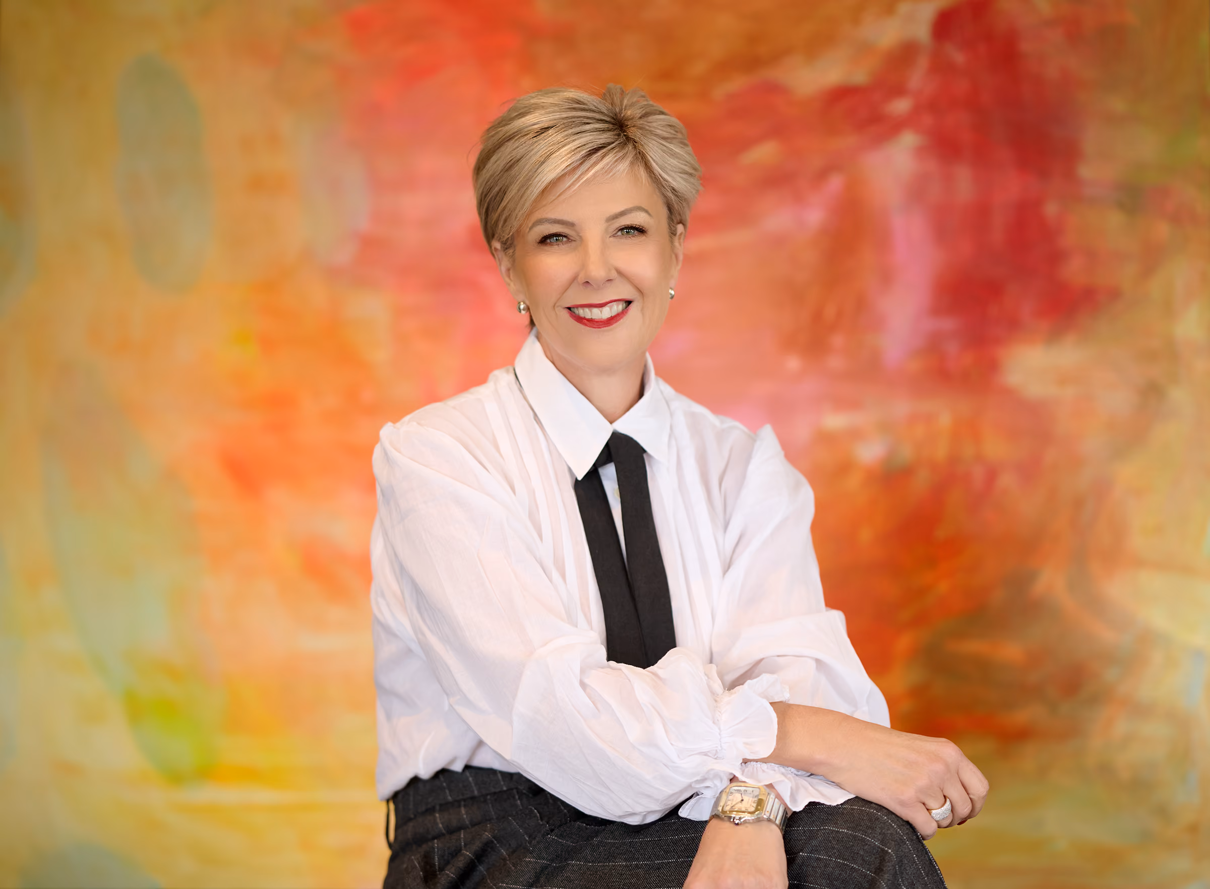 Nicole Moody, founder of Hunter Advisory in a white long sleeve shirt and black tie, sitting in front of a bright red, orange and yellow abstract painting