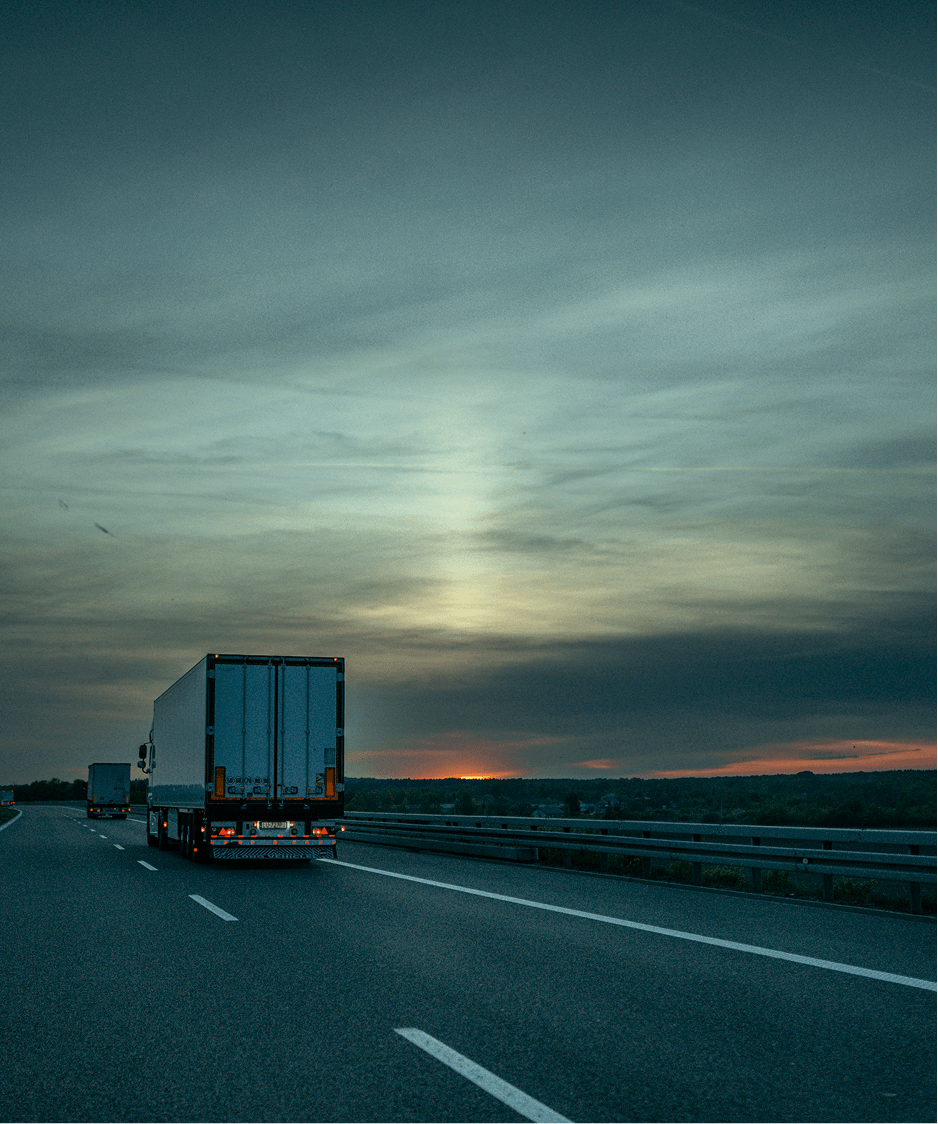 Due camion guidano su un'autostrada al tramonto con cielo nuvoloso e orizzonte visibile.