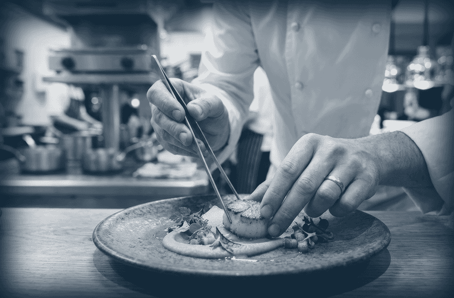 Un chef en veste blanche décore soigneusement une assiette gastronomique avec des pincettes.