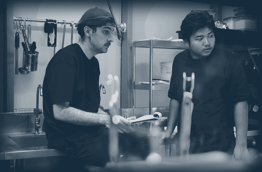 Deux hommes en tenue noire parlant dans une cuisine professionnelle.