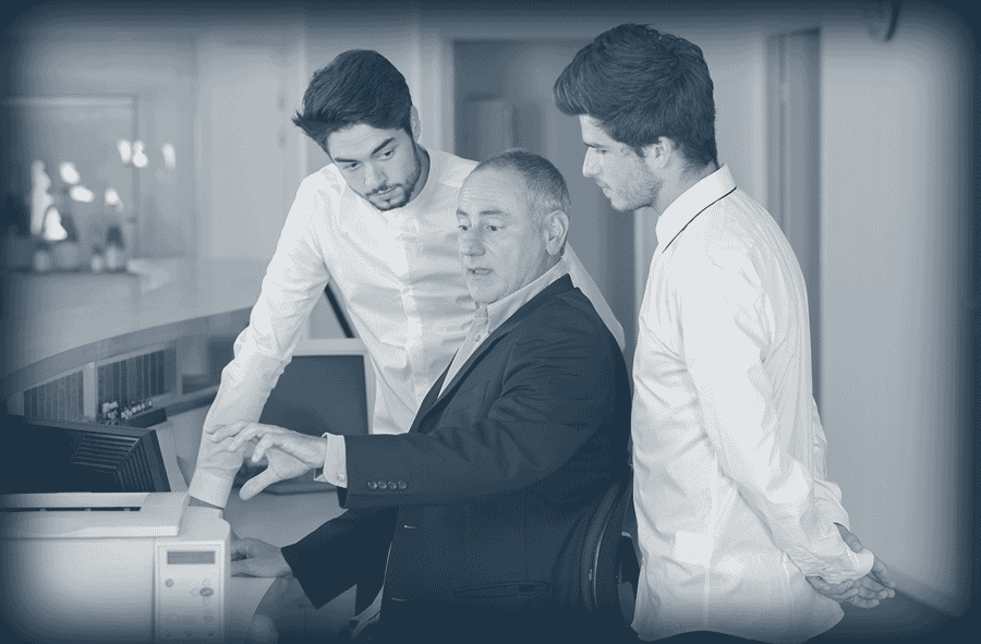 Un maître d'hotel explique quelque chose sur un écran d'ordinateur à deux jeunes hommes attentifs.