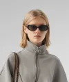 Blond woman wearing black sunglasses, gray jacket, and carrying a brown shoulder bag on light gray background.