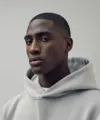 Portrait of a man wearing a light gray hoodie looking to the side against a plain background.