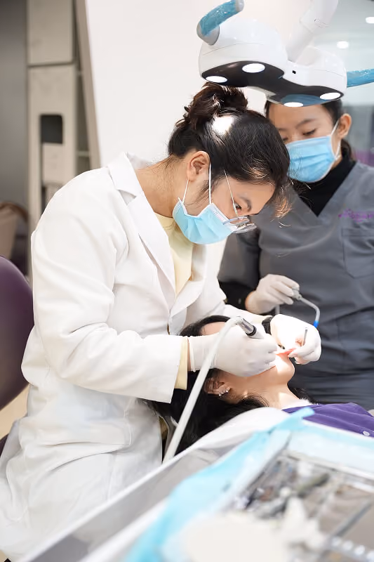 Dentist wearing mask and gloves treating a patient while assistant stands nearby in clinic.