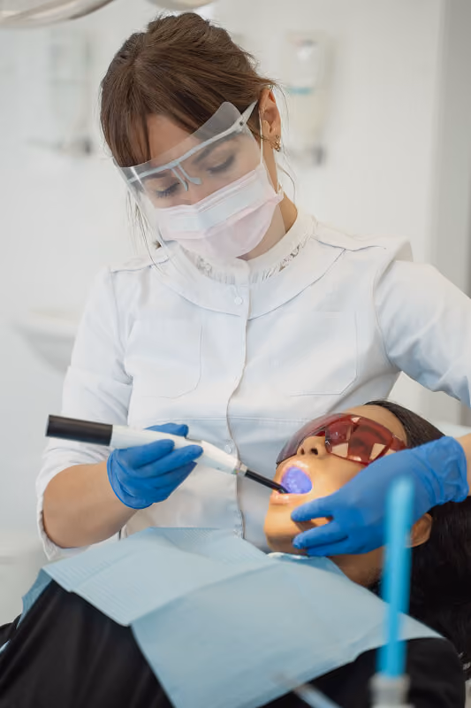 Dentist wearing mask and gloves uses a dental curing light on a patient wearing protective glasses.