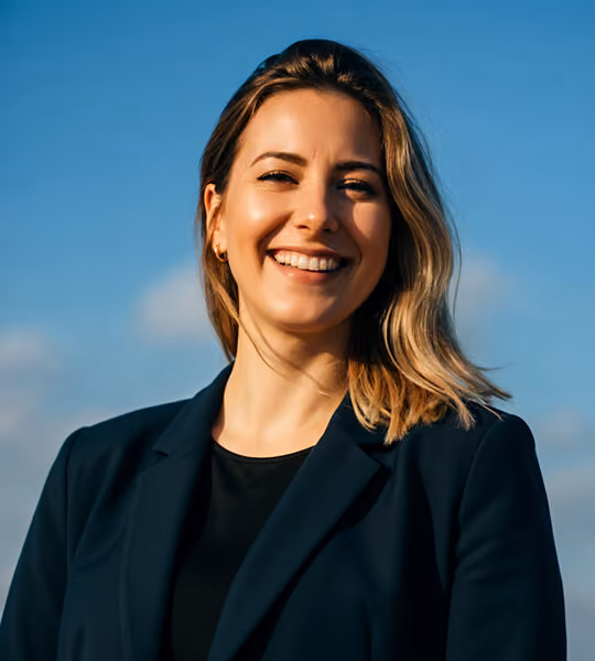 Smiling woman with blonde hair wearing a dark blazer against a blue sky background.