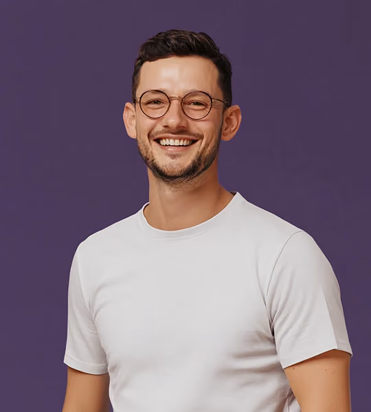 Smiling man with short dark hair, round glasses, and white T-shirt against a purple background.
