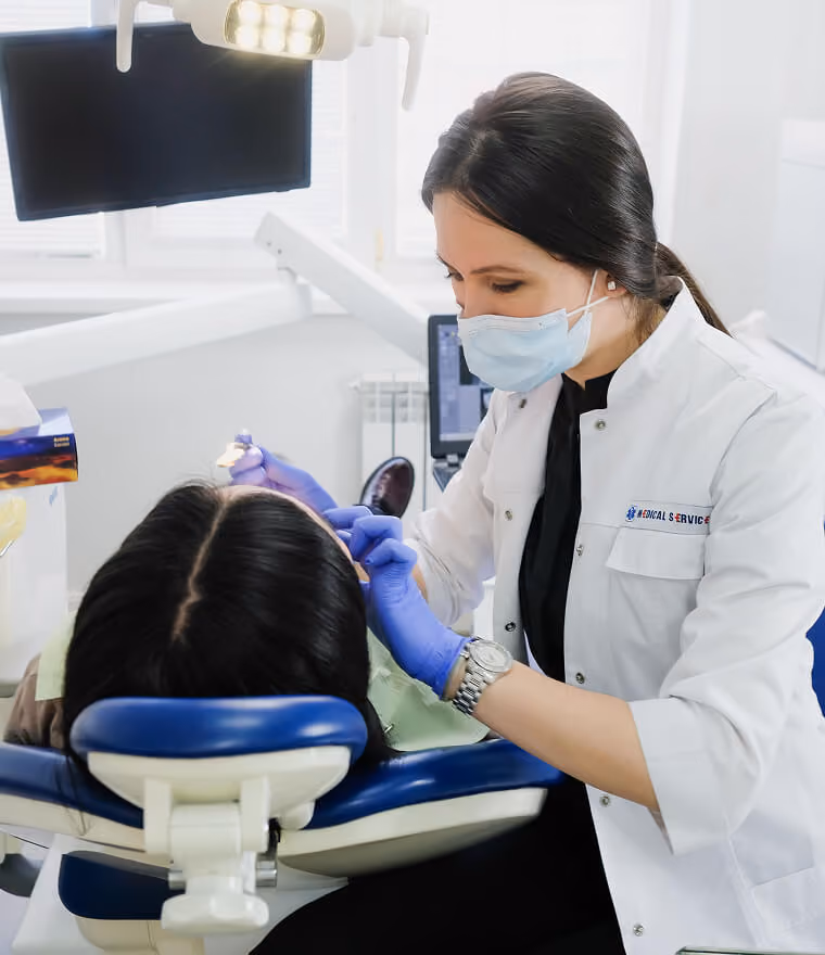 Dentist in mask and gloves examining a patient's teeth in a dental clinic chair.