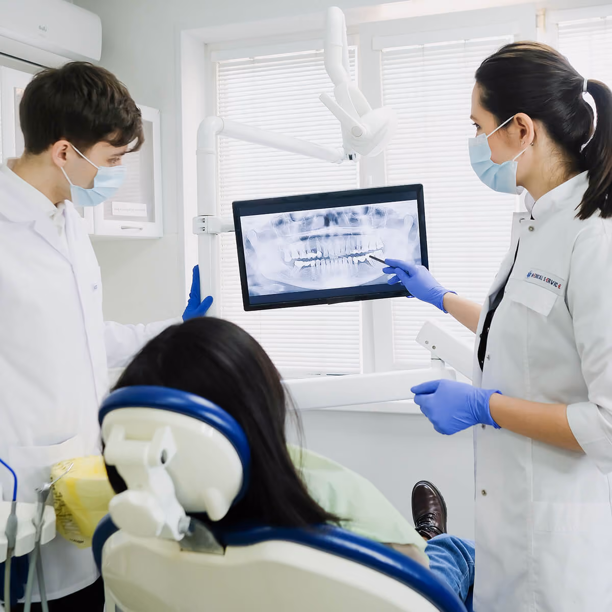 Two dentists with masks examine a dental X-ray on a monitor while a patient reclines in the chair.