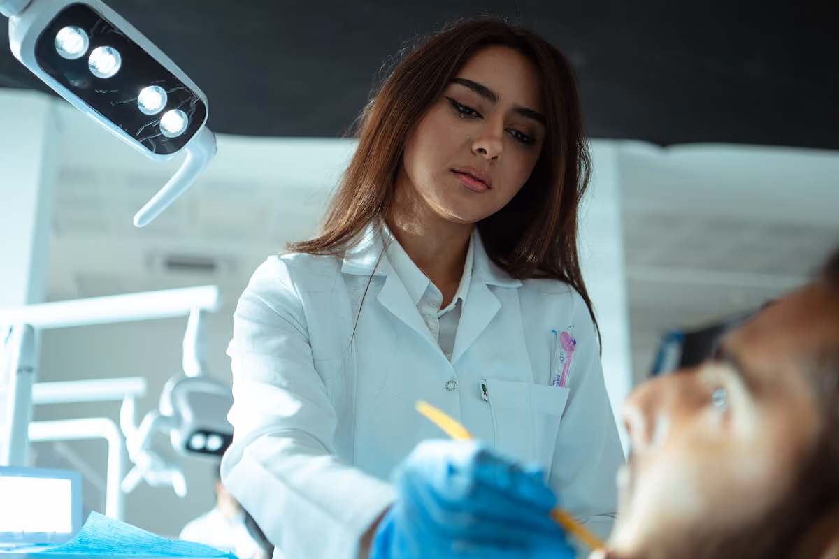 Female dentist in white coat with blue gloves working on a patient under dental lights.