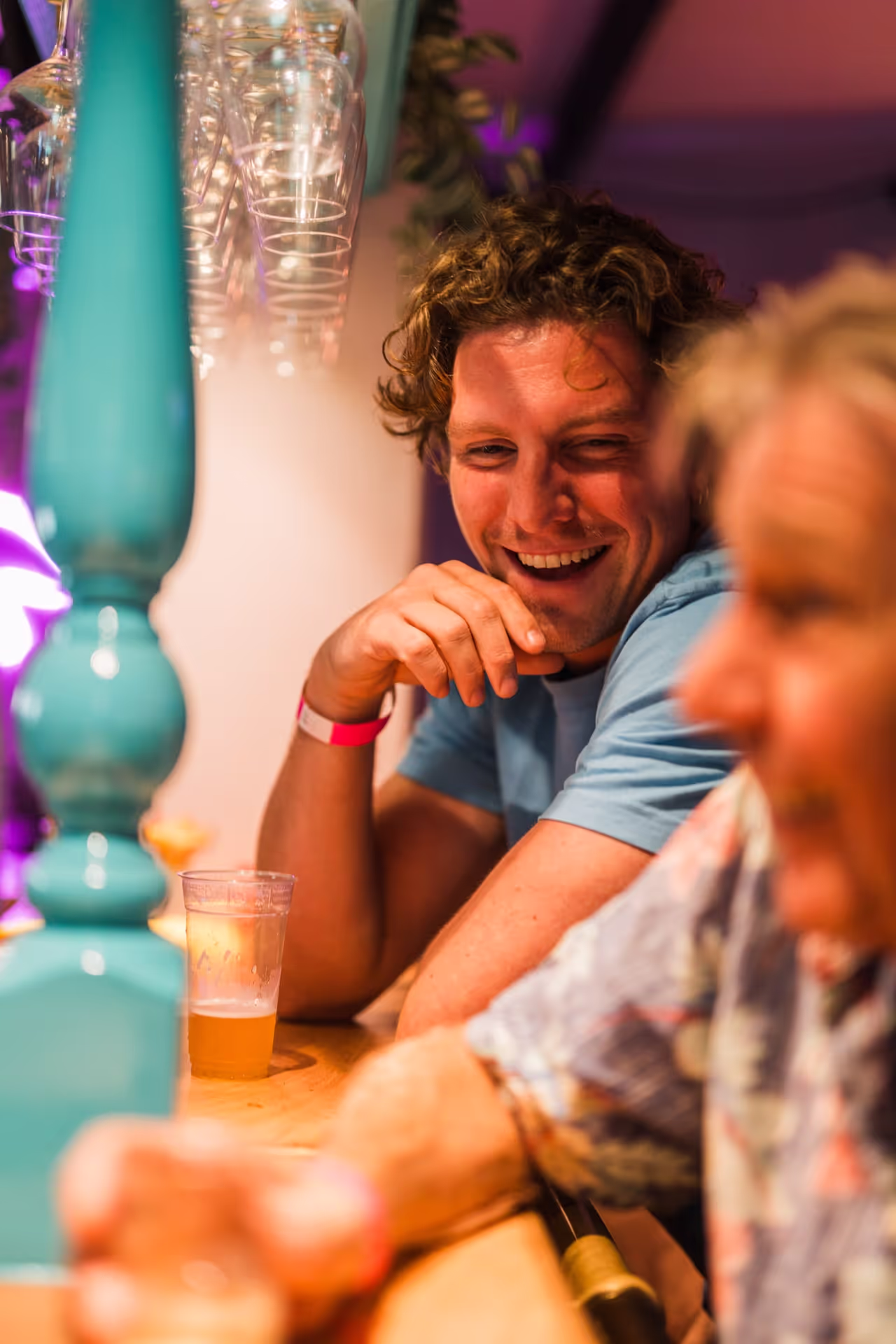 Man met blauwe blouse en roze polsbandje lacht tijdens een gezellige avond in de bar.