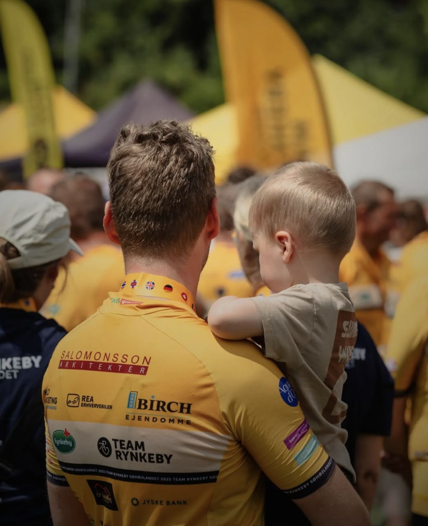 Team Rynkeby-syklister i gule trøyer, en med et barn på armen