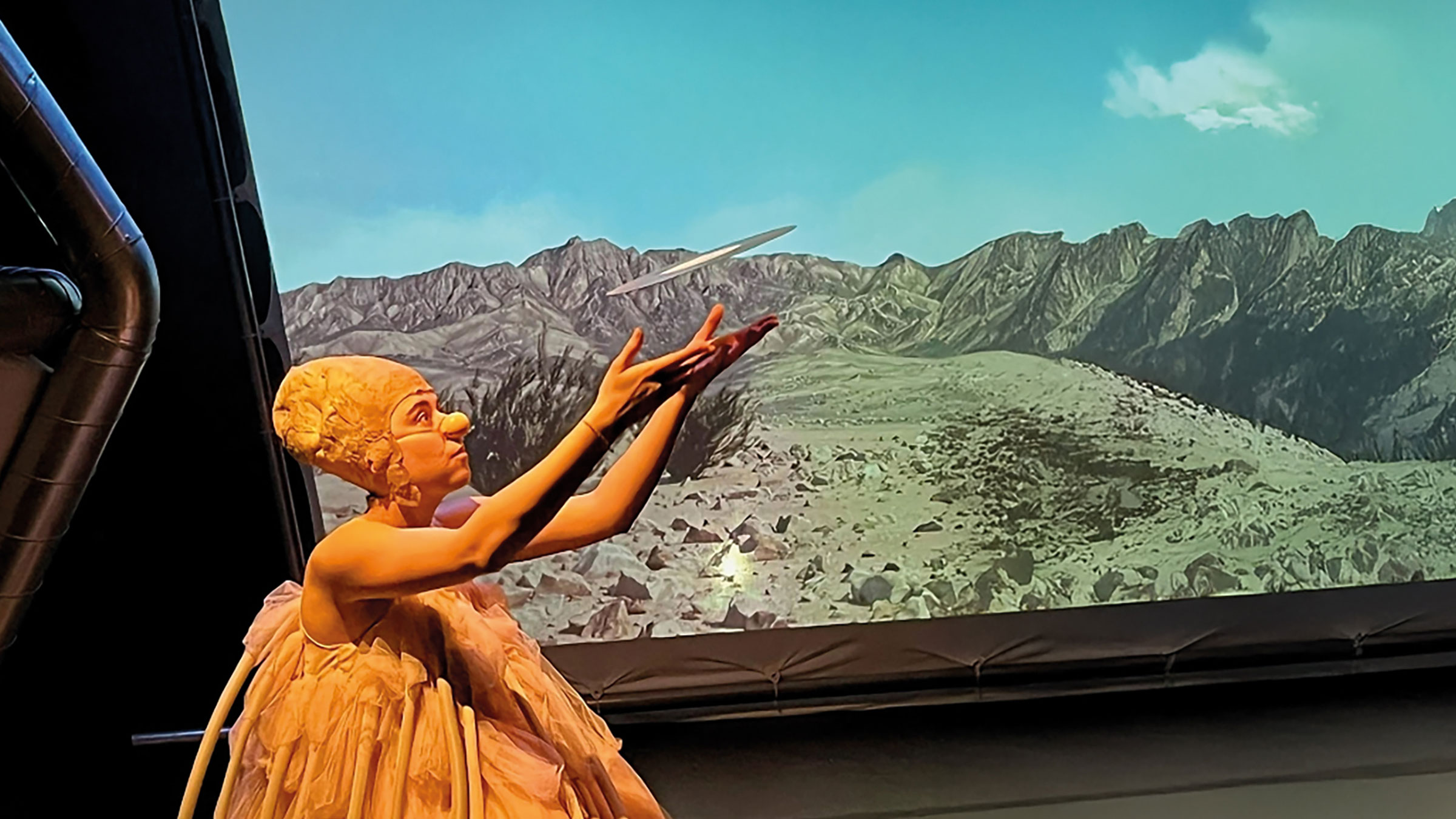 Aufführung im VISAVIS: Figur in goldenem Kleid vor einem gemalten Bergpanorama.