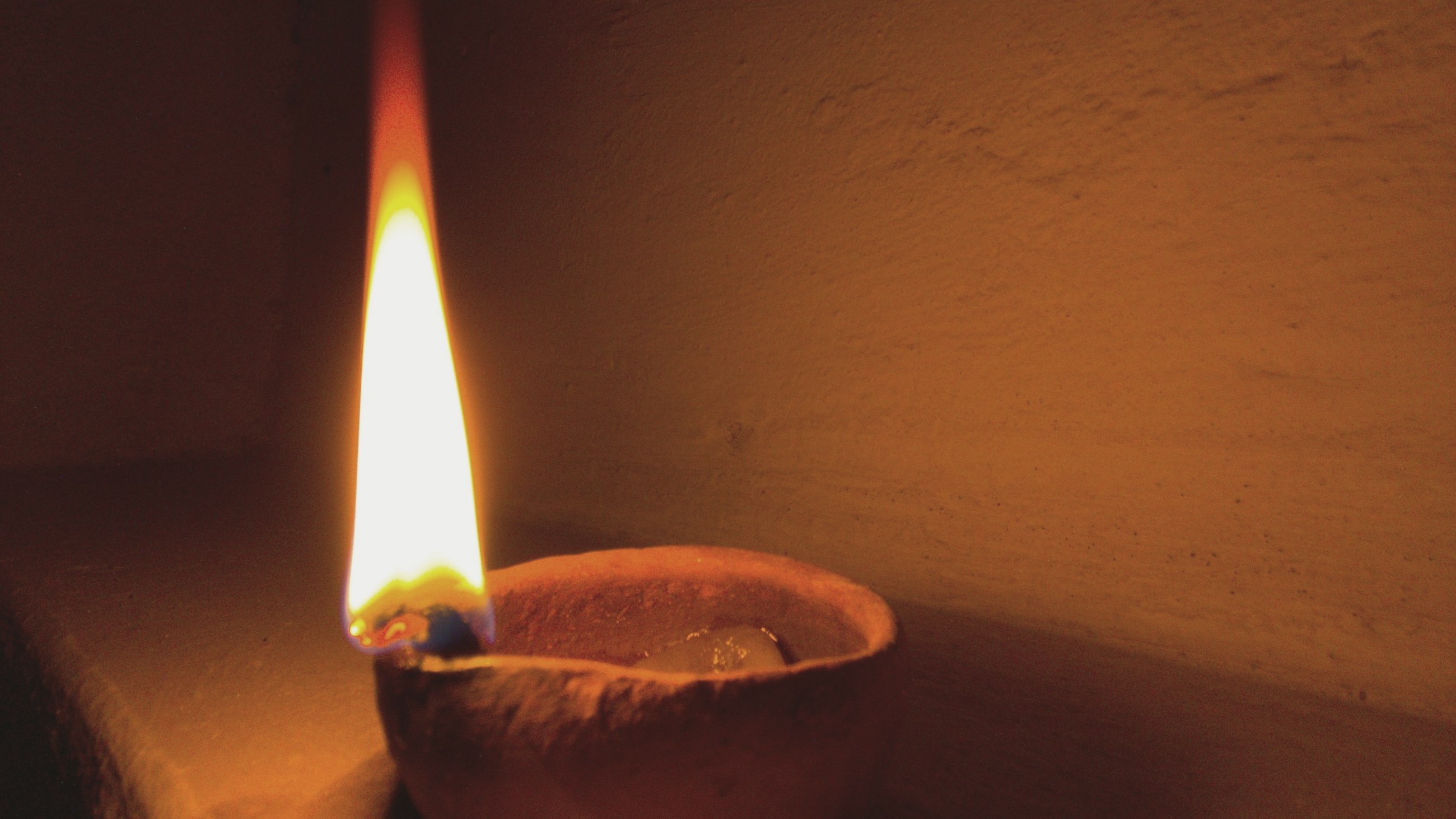 A single tall flame on a clay diya, burning against a dark wall.