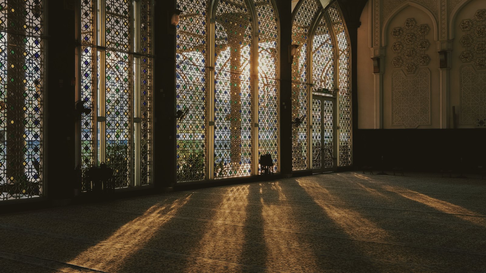 Long bands of daylight falling through tall latticed windows across an empty prayer hall.