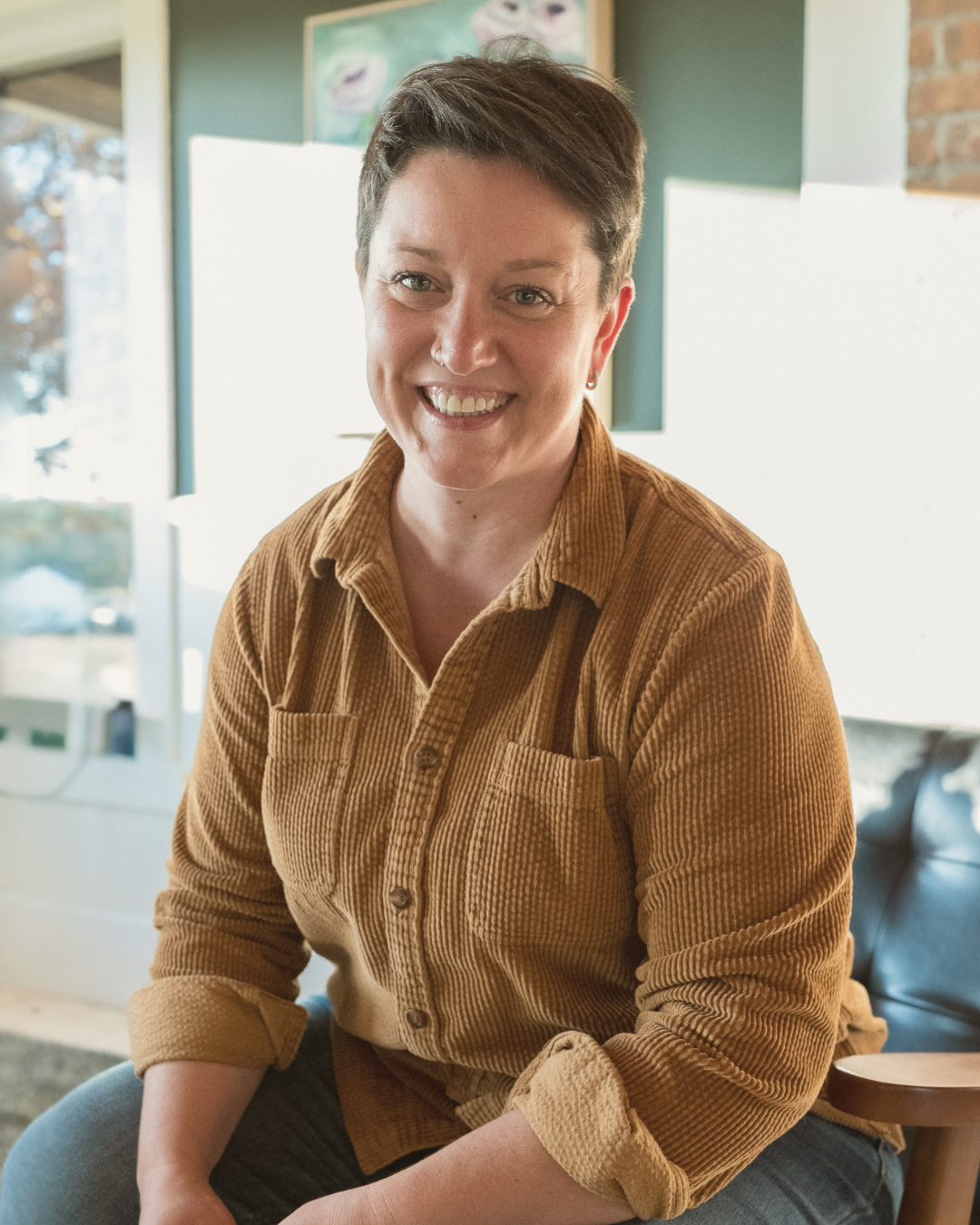 Keltie, smiling warmly, sits indoors in a well-lit room wearing a textured, mustard-colored button-up shirt. Her short hair and friendly expression add to her approachable presence. The background includes soft, neutral tones with hints of greenery and sunlight streaming in, giving the image a cozy and inviting feel.