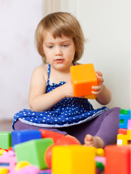 Toddler in a blue polka-dot dress playing with colorful toy blocks.
