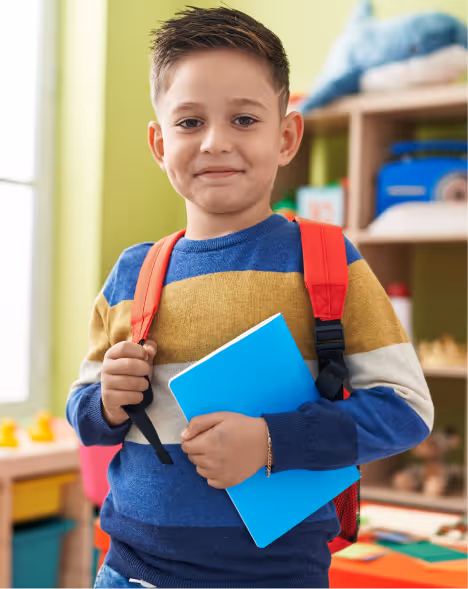 Smiling young boy wearing a striped sweater and red backpack holding a blue notebook in a colorful classroom.