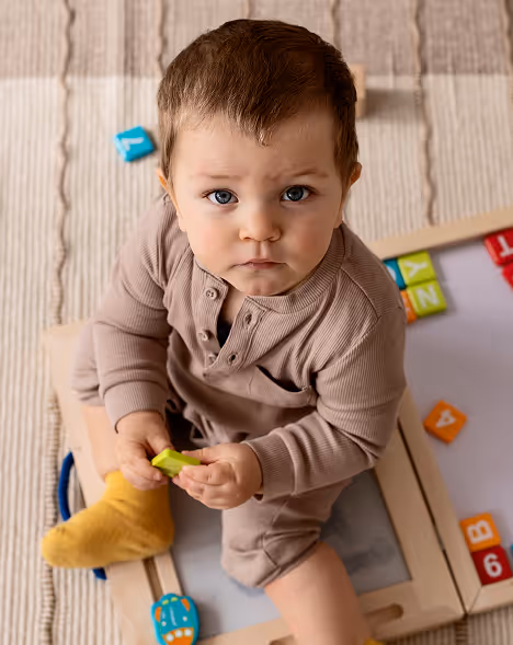 Toddler in beige outfit and yellow socks sitting on a wooden toy with colorful blocks scattered around.