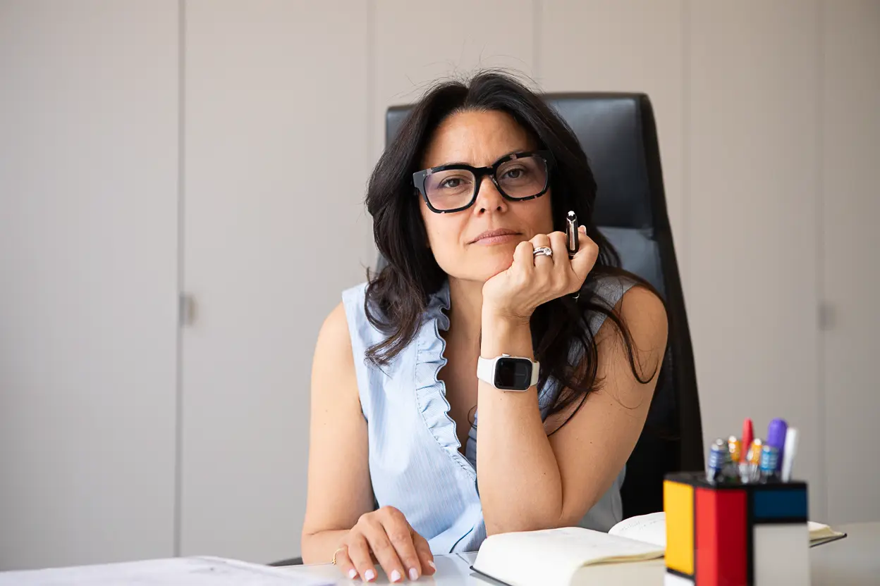 Donna seduta a una scrivania con espressione pensierosa, indossa occhiali, una camicia senza maniche azzurra e un orologio digitale bianco, con una mano che sorregge il mento e tiene una penna, davanti a lei un'agenda aperta e un portapenne colorato.