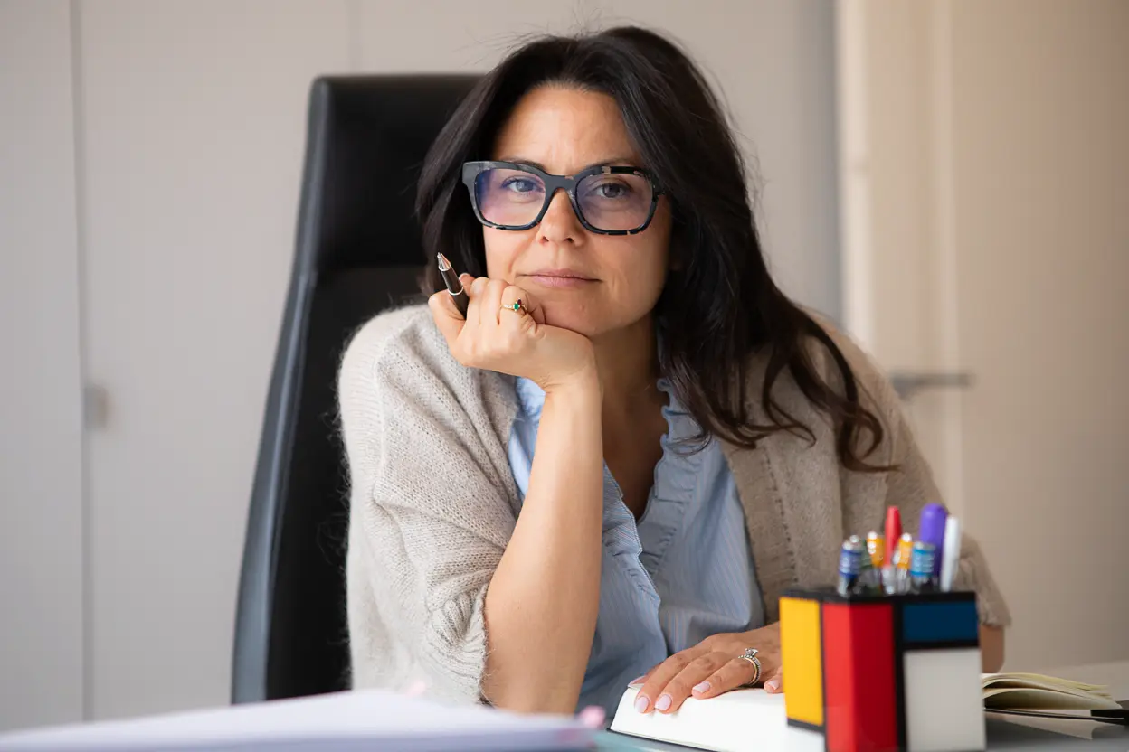 Donna con occhiali grandi e capelli scuri, seduta a una scrivania con un libro aperto e un portapenne colorato, che tiene una penna mentre guarda verso la fotocamera.
