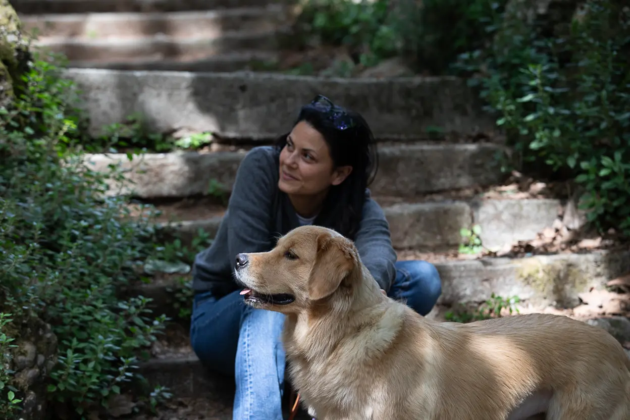 Donna con capelli scuri e maglione grigio seduta su gradini di pietra in un ambiente naturale, accanto a un cane di taglia media dal pelo dorato che guarda verso sinistra.