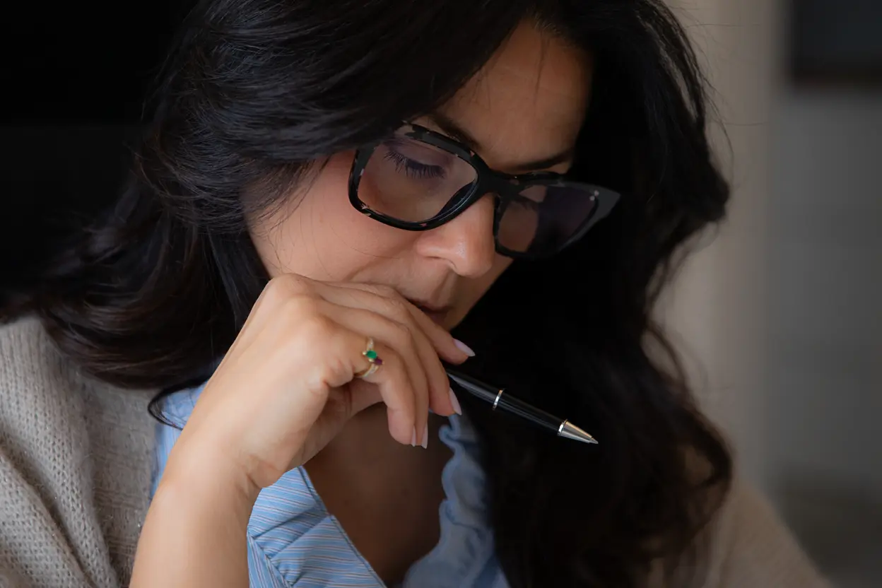 Donna con occhiali scuri che tiene una penna nera vicino alla bocca mentre guarda pensierosa verso il basso.